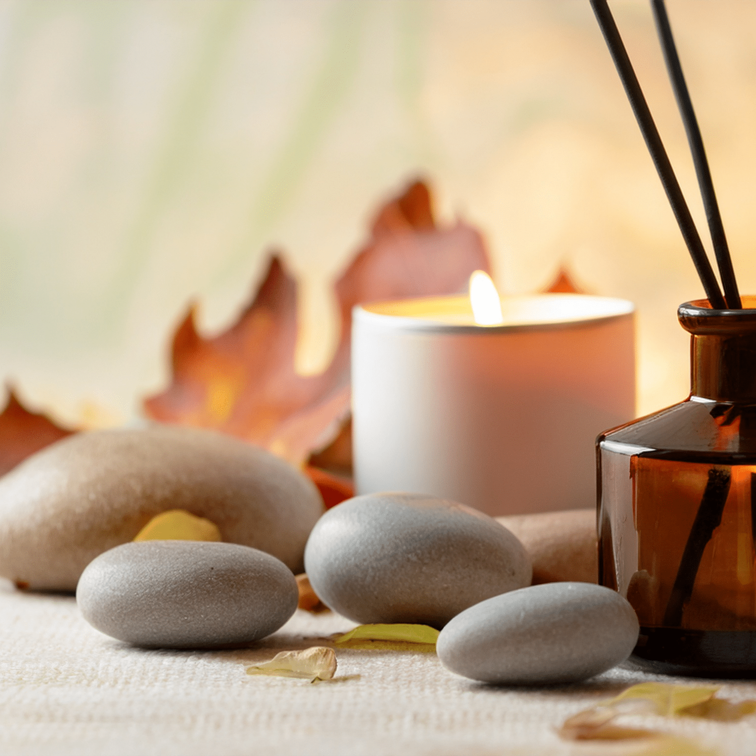 A collection of smooth stones arranged on a soft surface next to a white candle, an amber-colored bottle, and scattered autumn leaves, creating a calm and peaceful ambiance.