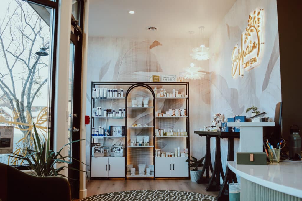 An interior view of a beauty spa featuring a display shelf filled with skincare products. A modern counter made of white material is visible, along with a decorative plant. The wall has a light-colored mural and illuminated signage.