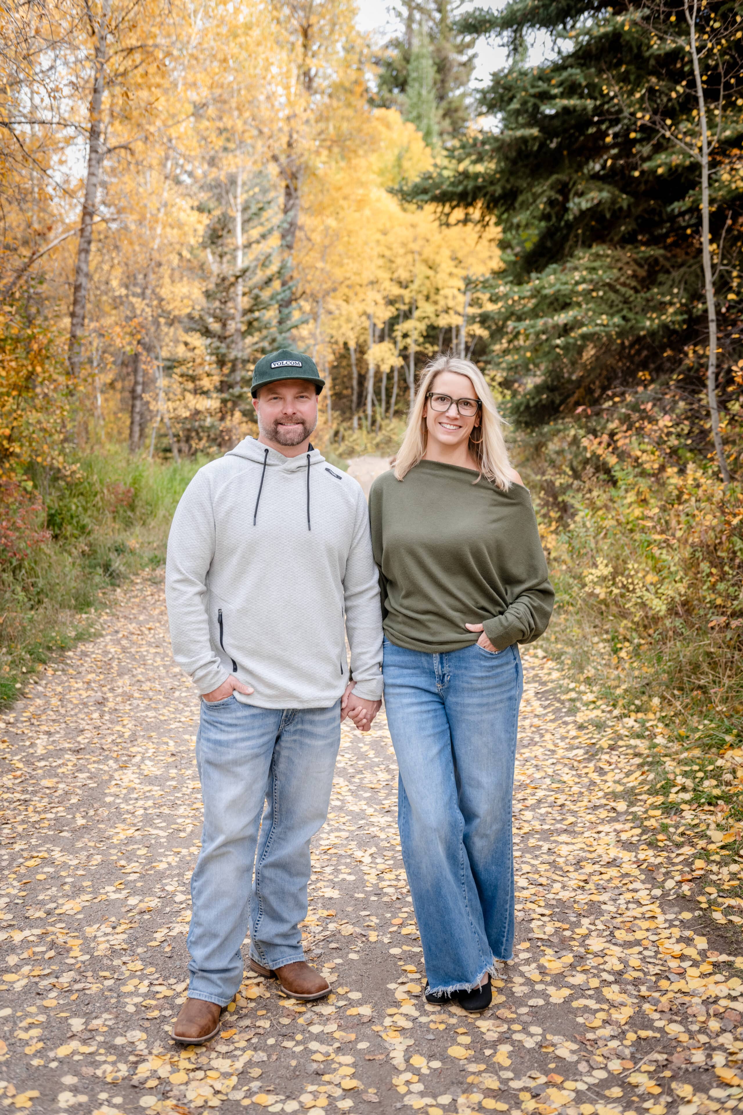 A man and a woman holding hands while walking down a path covered in yellow autumn leaves. The man wears a gray hoodie and a green cap, and the woman is dressed in a green sweater and wide-leg jeans. Background features trees with yellow foliage.