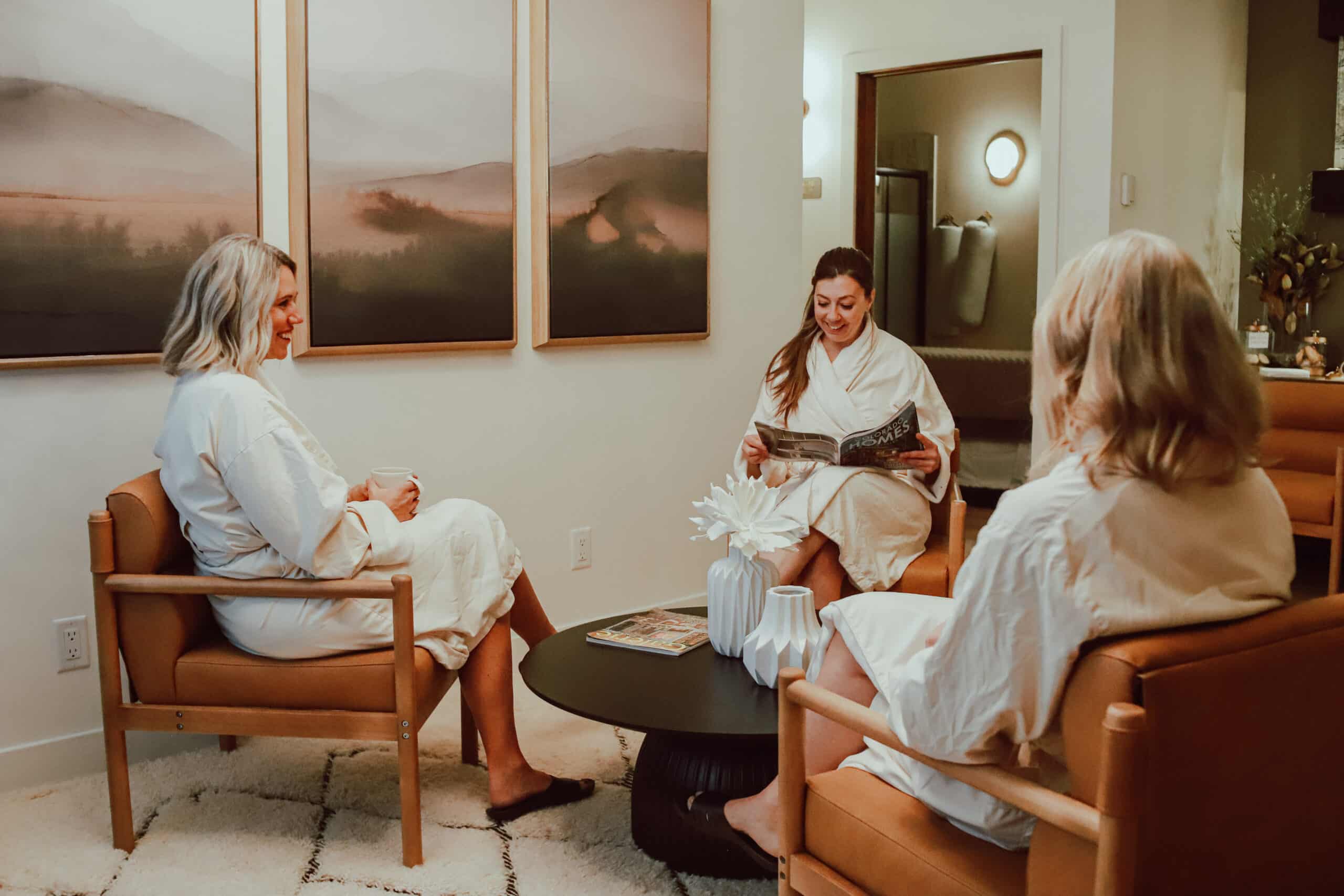 Three women in white bathrobes sitting in a cozy lounge area. One is reading a magazine, while the others engage in conversation. Framed landscape artwork hangs on the wall, enhancing the relaxing atmosphere.
