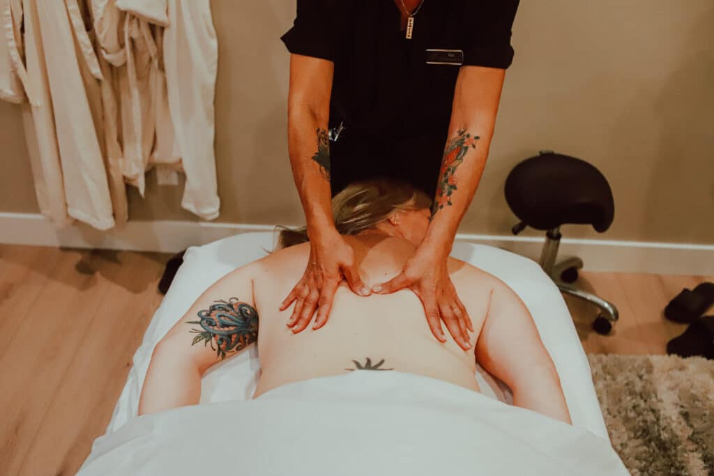 A massage therapist applying pressure to the back of a client during a massage session. The client has visible tattoos on their arms and is lying on a massage table covered with a white sheet.