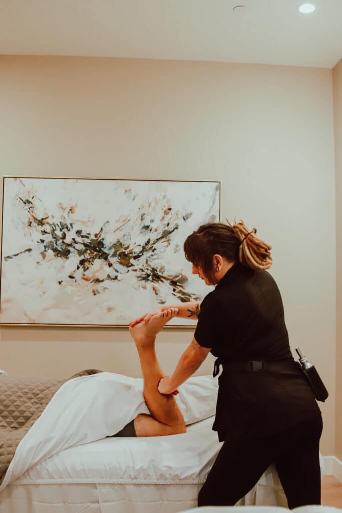 A massage therapist performing a leg stretch on a client lying on a treatment table, with a contemporary abstract painting hanging on the wall in a well-lit room.