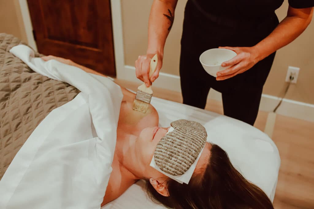A woman receiving a spa treatment lying on a massage table, with a therapist applying a product with a brush to her shoulders. The woman is wearing an eye mask.