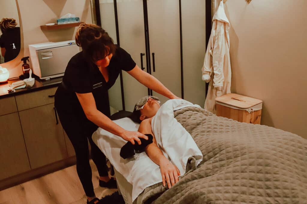 A spa therapist performing a massage on a client who is lying on a massage table, covered with a grey blanket. The treatment room is softly lit, with a cabinet and a robe hanging in the background.