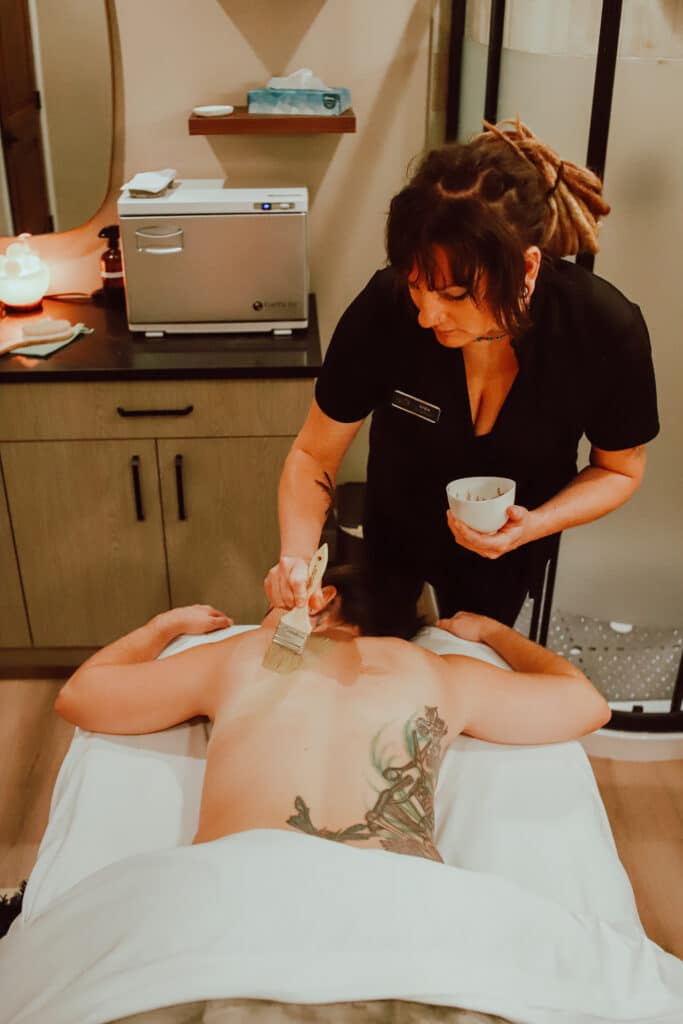 A spa setting where a massage therapist applies a treatment to a client's back. The client is lying face down on a treatment table, covered with a white sheet, and has a tattoo visible on their back.