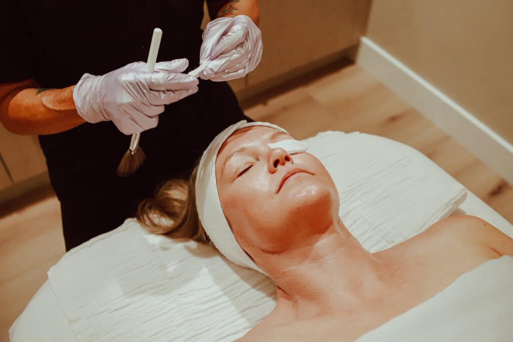 A woman lying on a treatment table with a headband, relaxed during a facial treatment. A technician, wearing gloves, is applying a product with a brush.