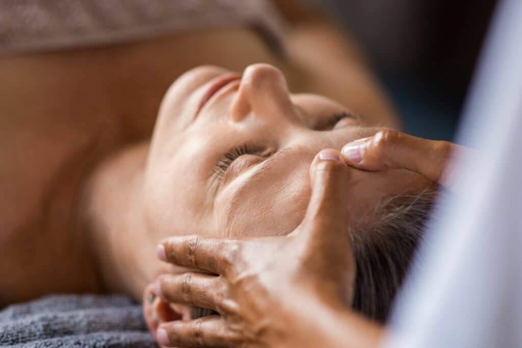 A close-up of a woman receiving a facial massage, with a therapist's hands gently pressing on her forehead. The setting appears calm and relaxing.