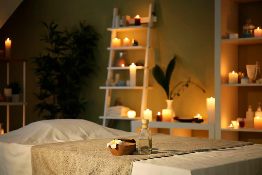 A tranquil spa room with a massage table covered in a beige cloth. A bowl of spa essentials and a glass bottle sit on the table. The room features shelves with candles and plants, creating a calming atmosphere.