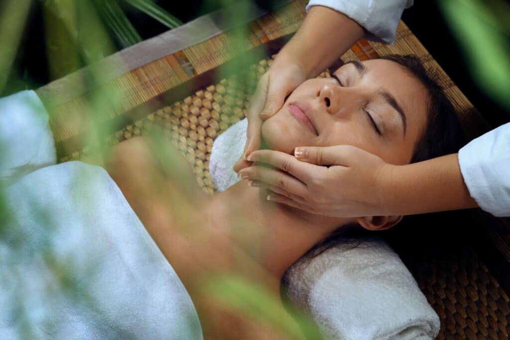 A woman receiving a facial massage while lying on a bamboo mat. She has her eyes closed and a relaxed expression, with a white towel draped around her shoulders. Green plants are visible in the foreground.