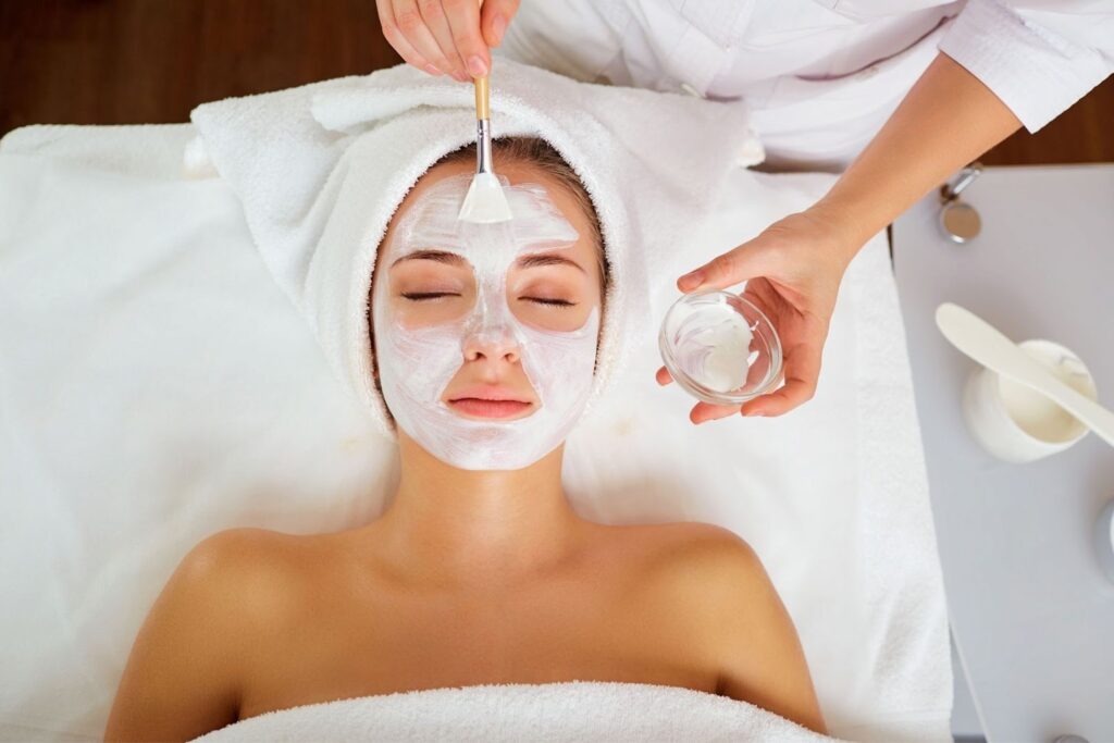 A woman receiving a facial treatment, lying on a treatment table covered with a towel. A skincare professional applies a mask to her face with a brush, while a small container of product is held nearby.