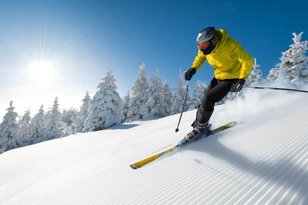 A skier in a bright yellow jacket descends a snowy slope, with snow-covered trees in the background and a bright sun shining in a clear blue sky.