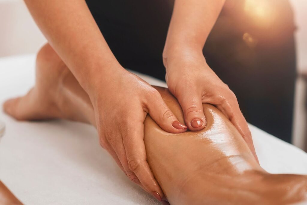 A person receiving a massage on their leg while lying on a massage table. The therapist's hands are applying pressure to the calf, and the leg appears moisturized. Soft lighting is present in the background, creating a calm atmosphere.