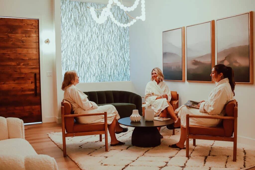Three women in bathrobes sitting in a serene lounge area with a modern design. They are engaged in conversation around a round black table. The room features neutral colors, a textured wall, and landscape artwork on display.