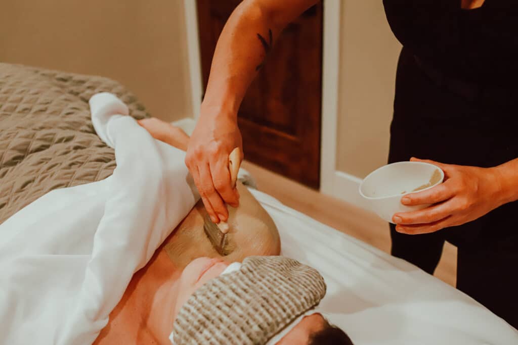 A person receiving a massage treatment while lying on a bed covered with white linens. The therapist applies a clay mask to the client's shoulder with a brush, and a small bowl is nearby. This is a spa experience in Steamboat Springs.