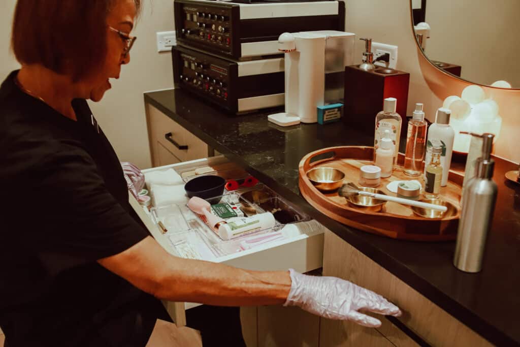 A woman wearing a black shirt and gloves organizes items at a wooden counter. She is pulling open a drawer that reveals various tools and products, including bottles and containers, while a tray with additional items is positioned on the counter's surface.