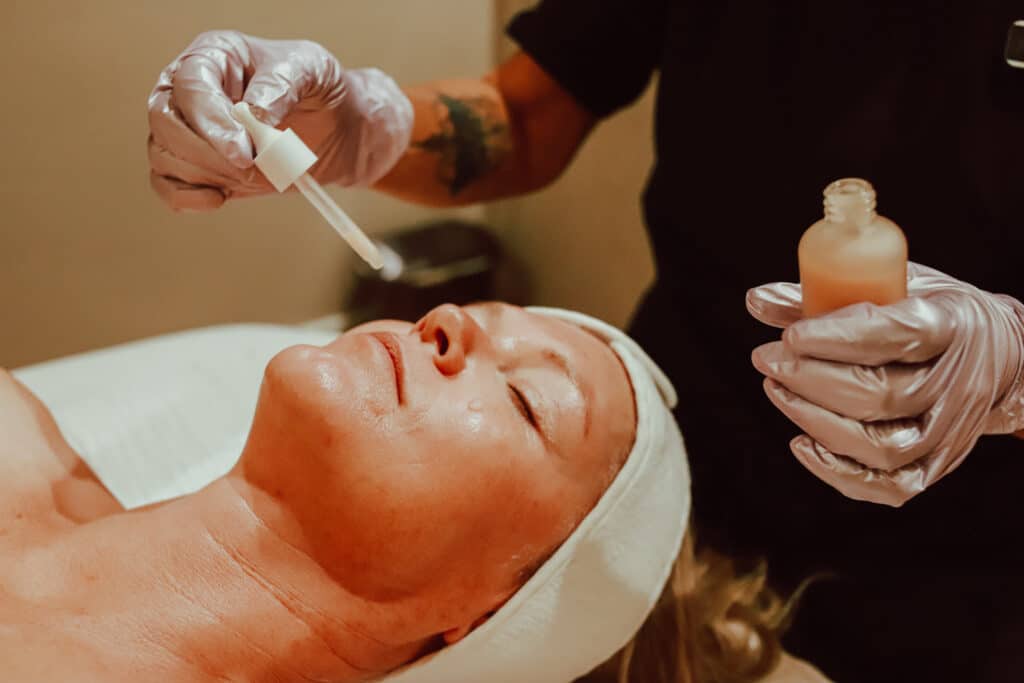 A person receiving a facial treatment, lying on a treatment bed with a white headband. A technician in gloves applies a dropper of serum to the client's face, while holding a small bottle of another product. The setting appears calm and professional.