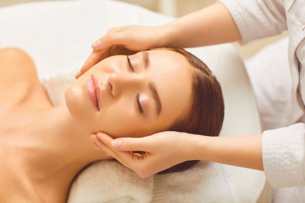A woman receiving a facial treatment at a spa, her eyes closed and relaxed. A massage therapist is gently holding her face, applying pressure as part of the treatment. The background is softly blurred, highlighting the serene environment.