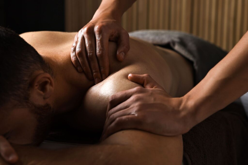 A person receiving a massage lies face down on a treatment table. One therapist uses both hands to apply pressure on the person's shoulders and upper back. The setting appears tranquil with soft lighting and a neutral color palette.