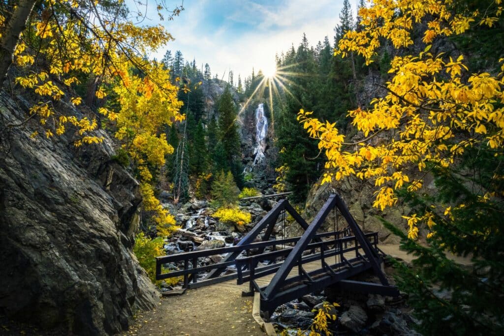 A narrow path leads through a forest with vibrant yellow leaves. A wooden bridge crosses over rocky terrain, and a waterfall cascades in the background. Sunlight beams through the trees, casting a warm glow across the scene.