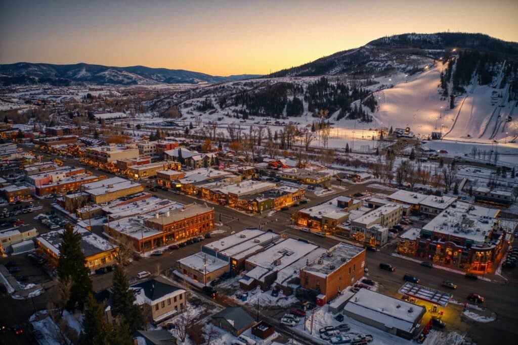 An aerial view of a snow-covered town at sunset, featuring illuminated buildings lining the streets. Snow-capped hills are visible in the background, with a ski slope on the right. Warm light highlights the buildings, creating a cozy atmosphere.