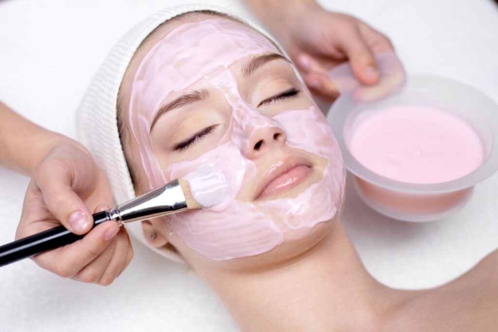 A woman receiving a facial treatment, with a light pink mask applied on her face. One hand holds a brush applying more product, while a bowl of the same mask is in the foreground. The setting appears to be a spa or beauty treatment room.