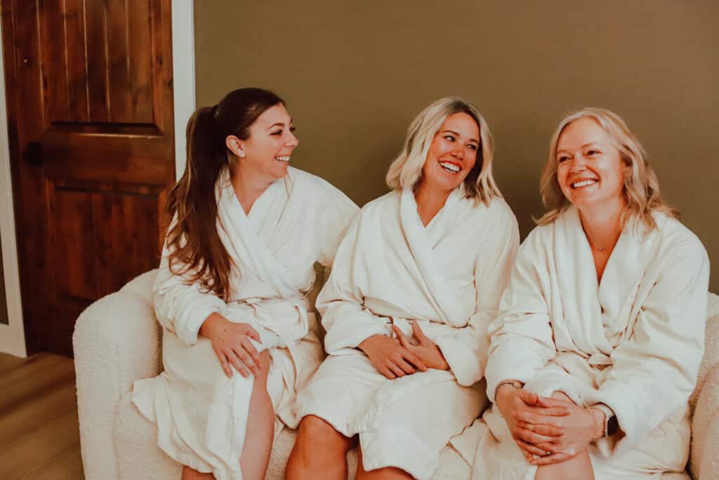 Three women wearing white bathrobes are seated on a plush white sofa. They are positioned closely together against a green wall, with one woman on the left and two women on the right, all appearing engaged in conversation.
