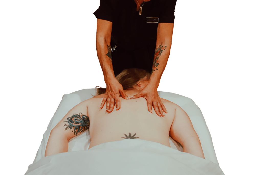 A person receiving a massage lies face down on a treatment table, covered with a white sheet. A massage therapist, wearing a black shirt, applies pressure to the client's back with both hands. Visible tattoos are on the client's arms.