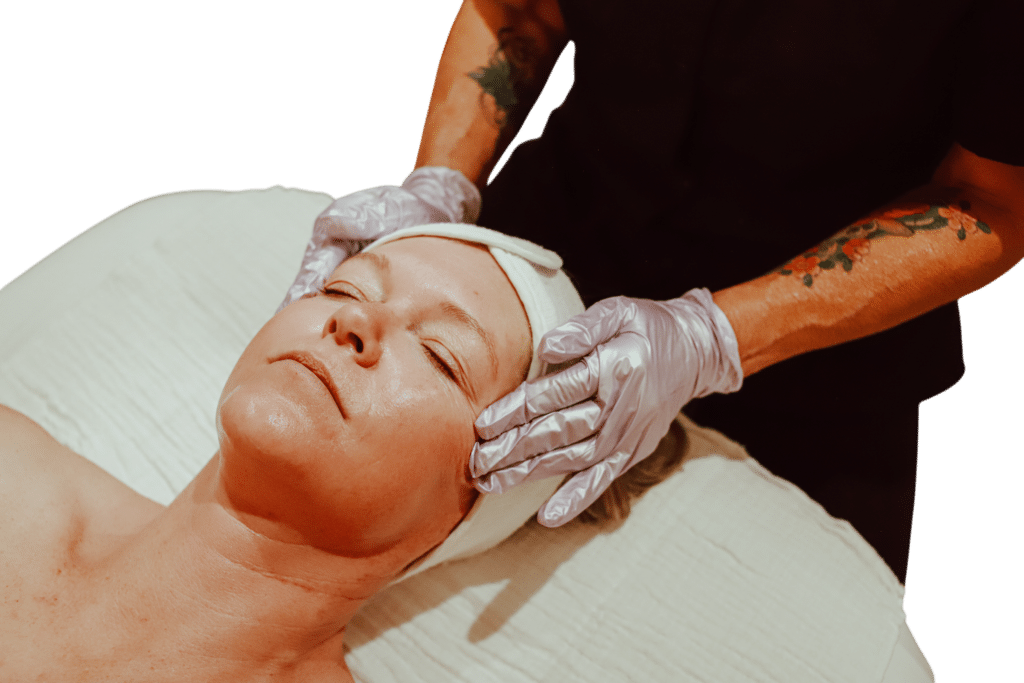 A person receiving a facial treatment while lying on a treatment table. An individual with gloved hands is gently massaging the client's face, who has her eyes closed and is wearing a white headband. The background is blurred.