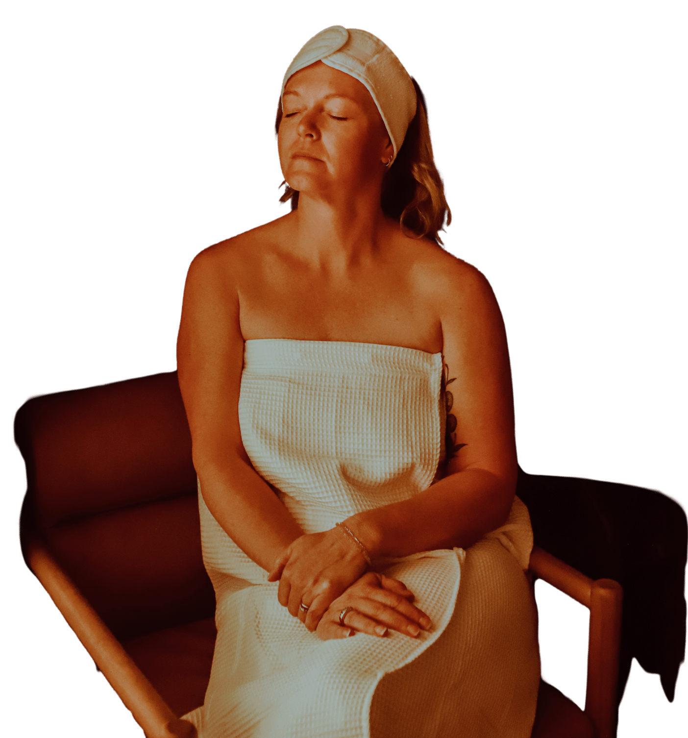A woman with blonde hair, wearing a white towel and a matching headband, sits on a brown chair with her eyes closed. The background is dark, highlighting her relaxed posture and the simple, elegant setting.
