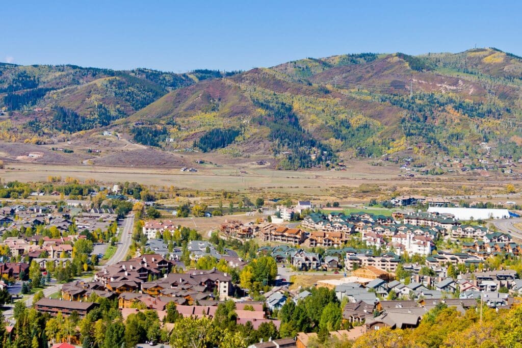 A mountain town with clustered houses and roads in the foreground, set below tree-covered hills under a clear blue sky.