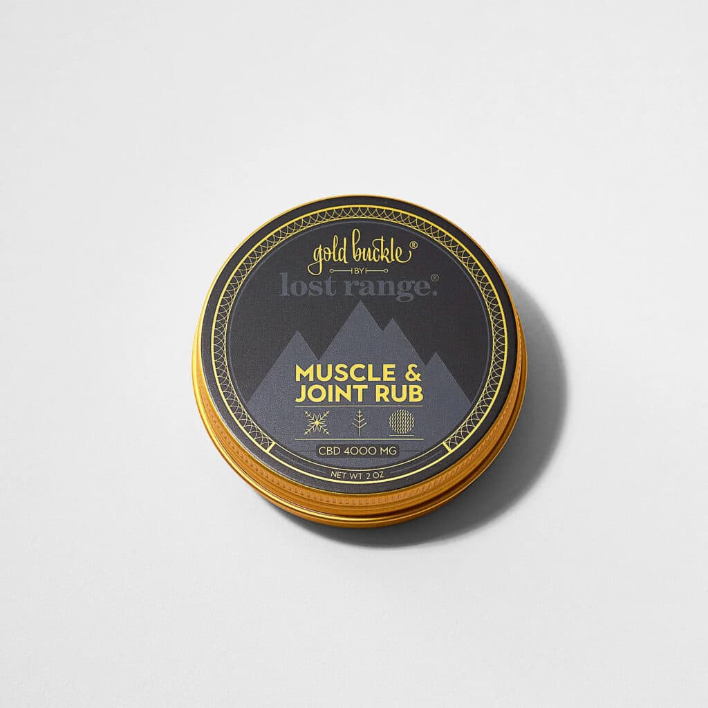 A round gold tin of Gold Buckle Lost Range Muscle & Joint Rub sits on a light gray surface with a soft shadow.