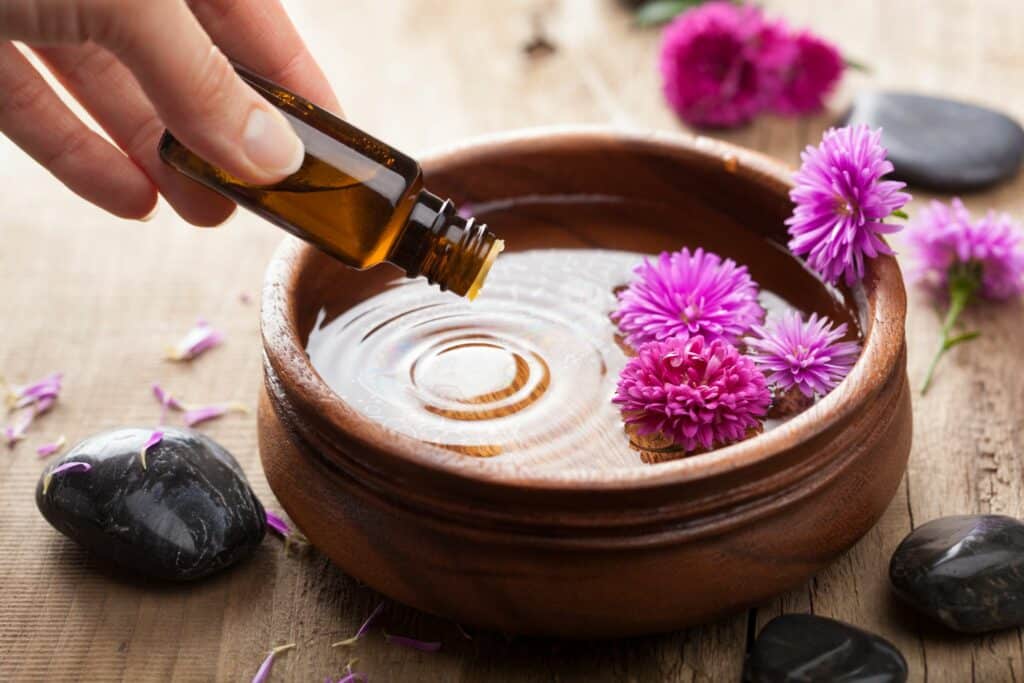 A hand pours amber oil into a wooden bowl of water beside purple flowers and black stones on a rustic table.