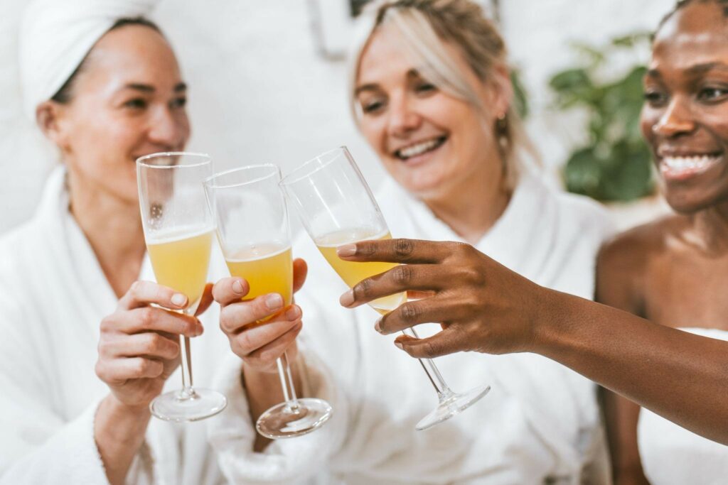 Three women in white robes clink champagne flutes filled with orange juice in a bright room.