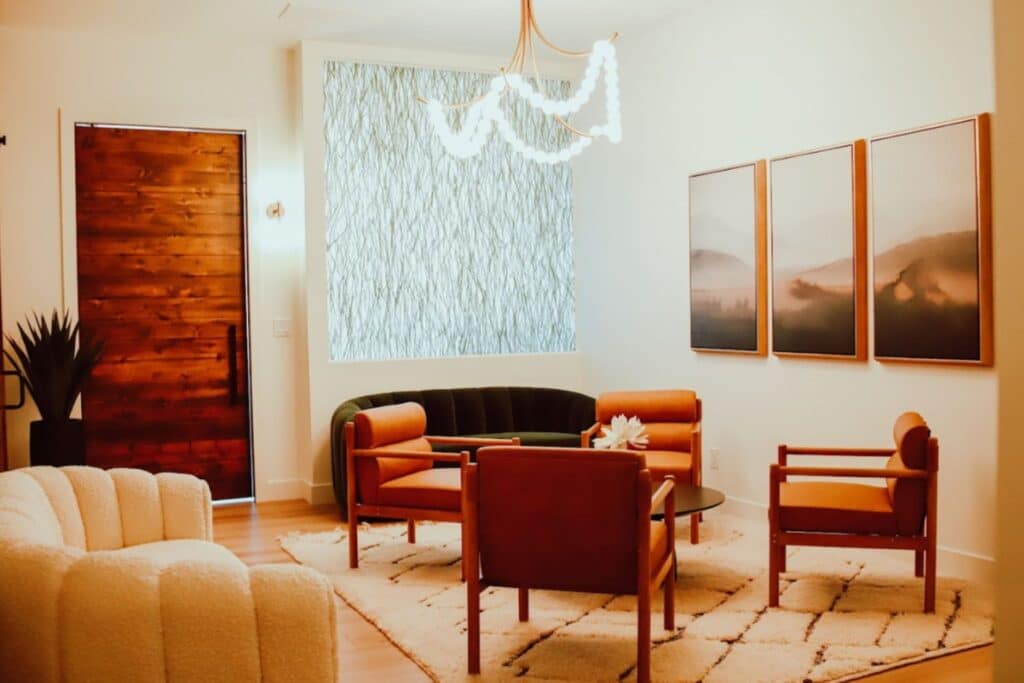 A modern living room with wooden armchairs around a circular coffee table, a beige sofa, and framed landscape art on white walls.