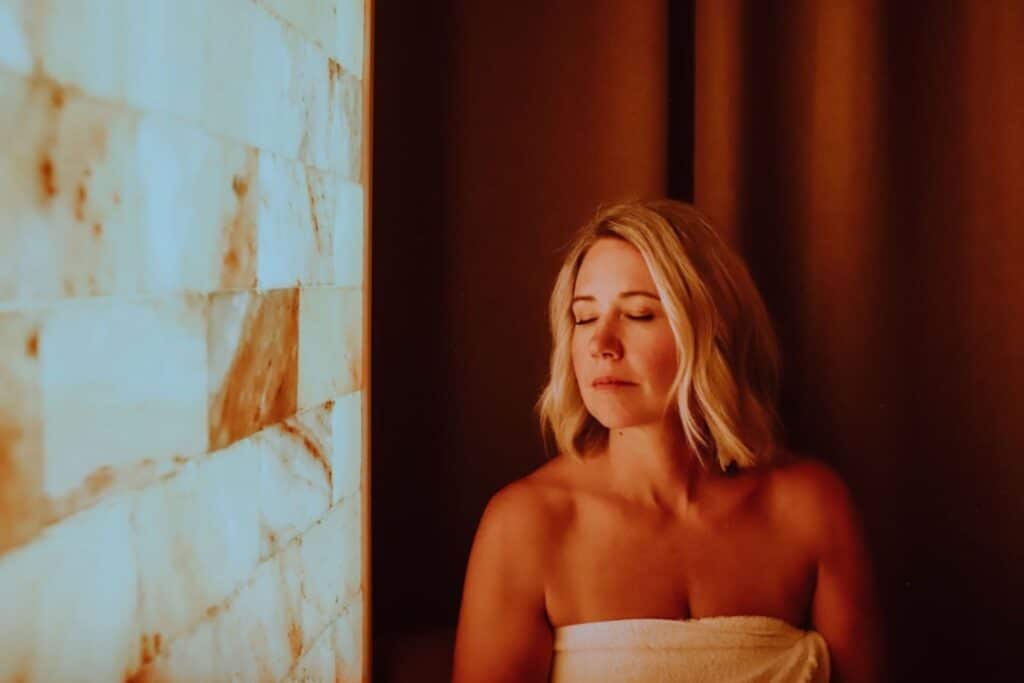 A blonde woman with a towel wrapped around her chest sits beside a glowing salt wall in a dim spa room.