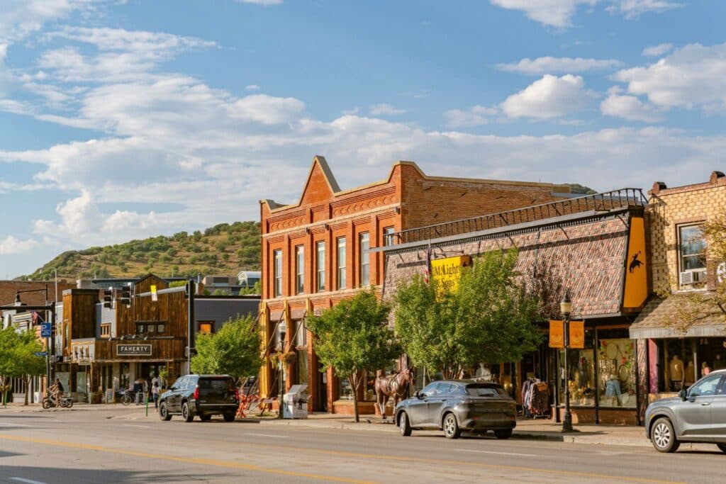 Historic downtown street with red-brick storefronts, parked cars, and trees lining the sidewalk under a blue sky with scattered clouds.